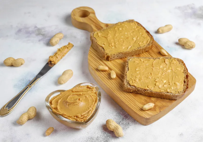 sanduíches de manteiga de amendoim em cima da tabua de madeira com um pode em formato de coração com manteiga de amendoim com uma faca do lado 