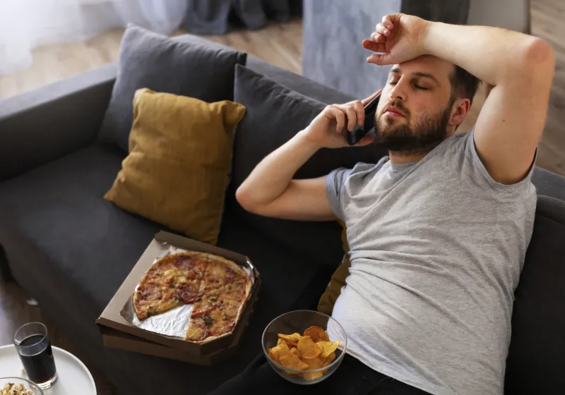 homem deitado no sofá passando mal com mão na cabeça e comendo junk food com smartfone na mão e a mão na cabeça 