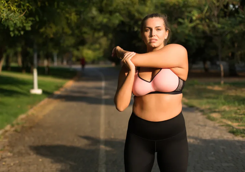 linda garota plus size em top esportivo rosa e leggings fazendo alongamento no parque 