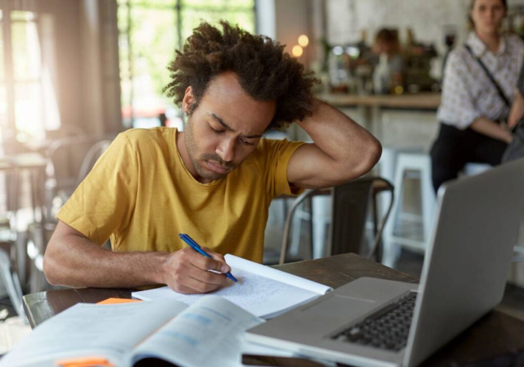 homem afro americano tentando se concentra sentado no cafe escrever em seu caderno com computador aberto cocando a cabeça