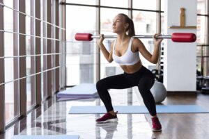 Imagem de uma modelo fazendo exercício para glúteos com uma barra em seus ombros dentro de uma academia de dia