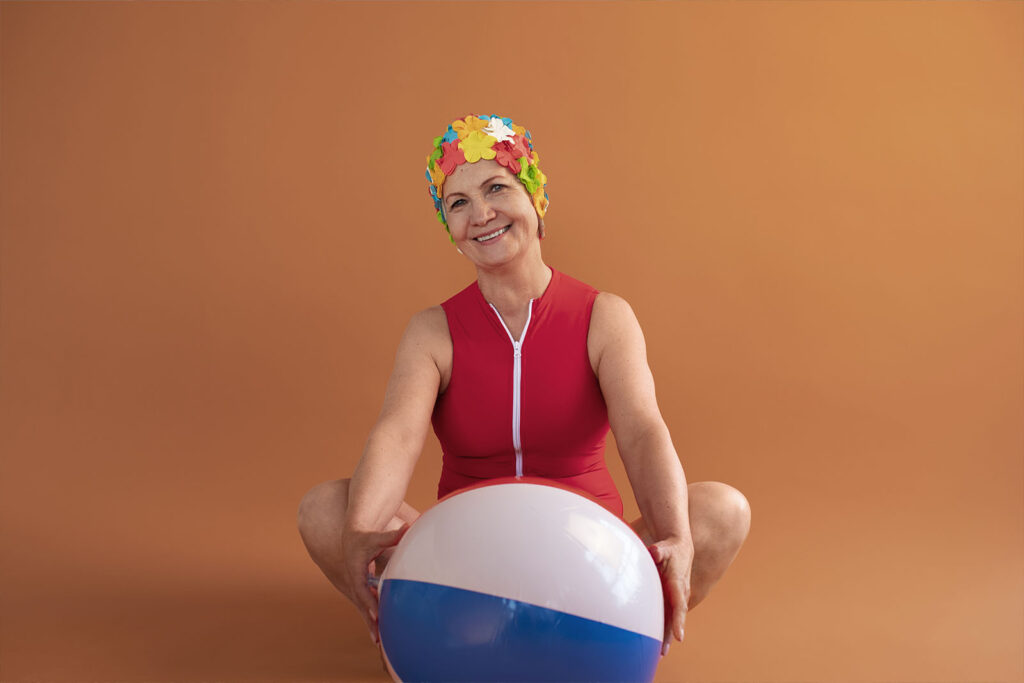 Imagem de uma senhora de idade sentada no chão, vestindo um maiô vermelho e touca florida para piscina, com uma bola de hidroginástica em sua frente, com o fundo de estúdio laranja 