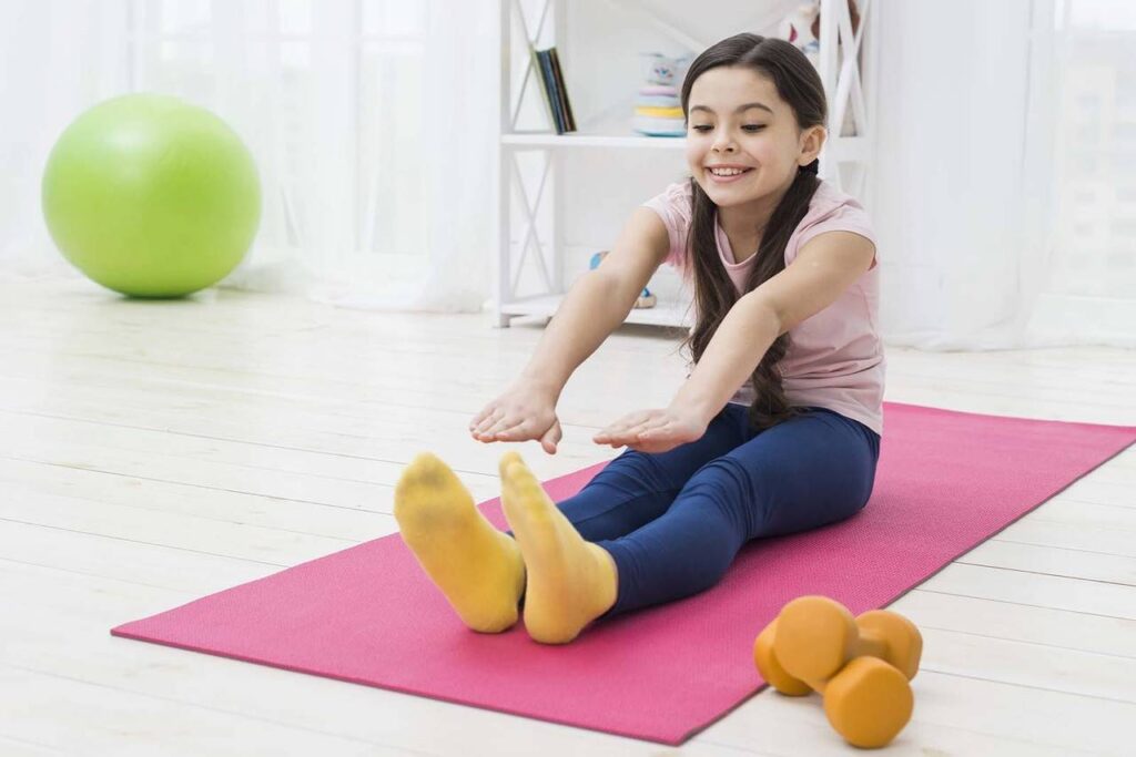 Imagem de uma menina sentada em um tapete rosa no chão, vestindo roupas de praticar exercício, fazendo alongamento, com o fundo de sala de estar 