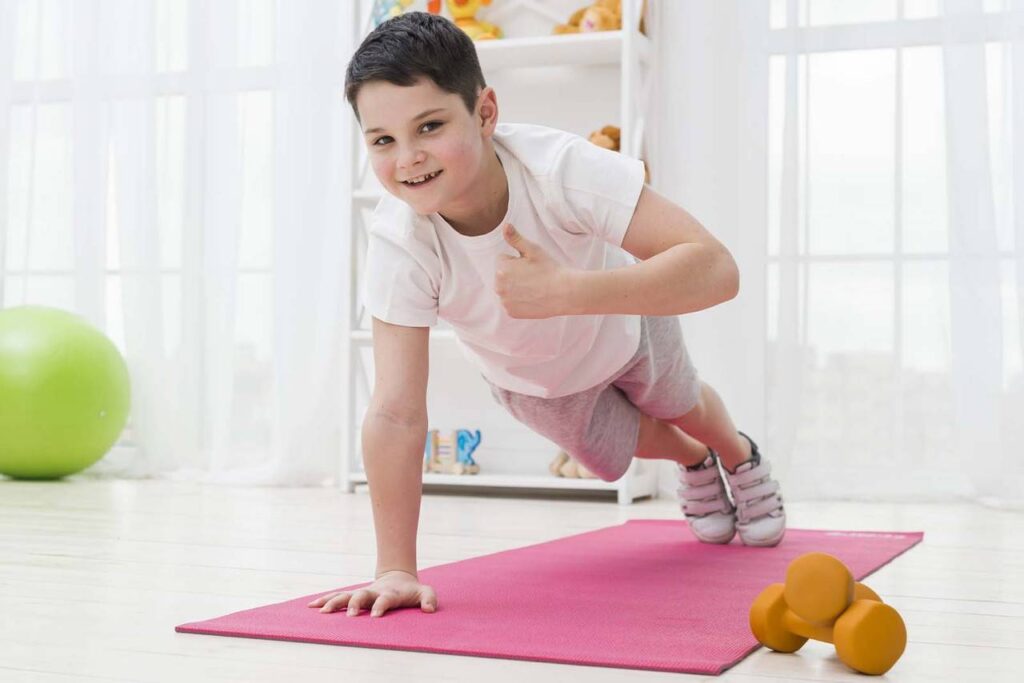 Imagem de um menino fazendo o exercício de prancha, em cima de um tapete rosa no chão, com uma das mãos fazendo um jóia, vestindo roupas de fazer exercício, com o fundo de sala de estar  