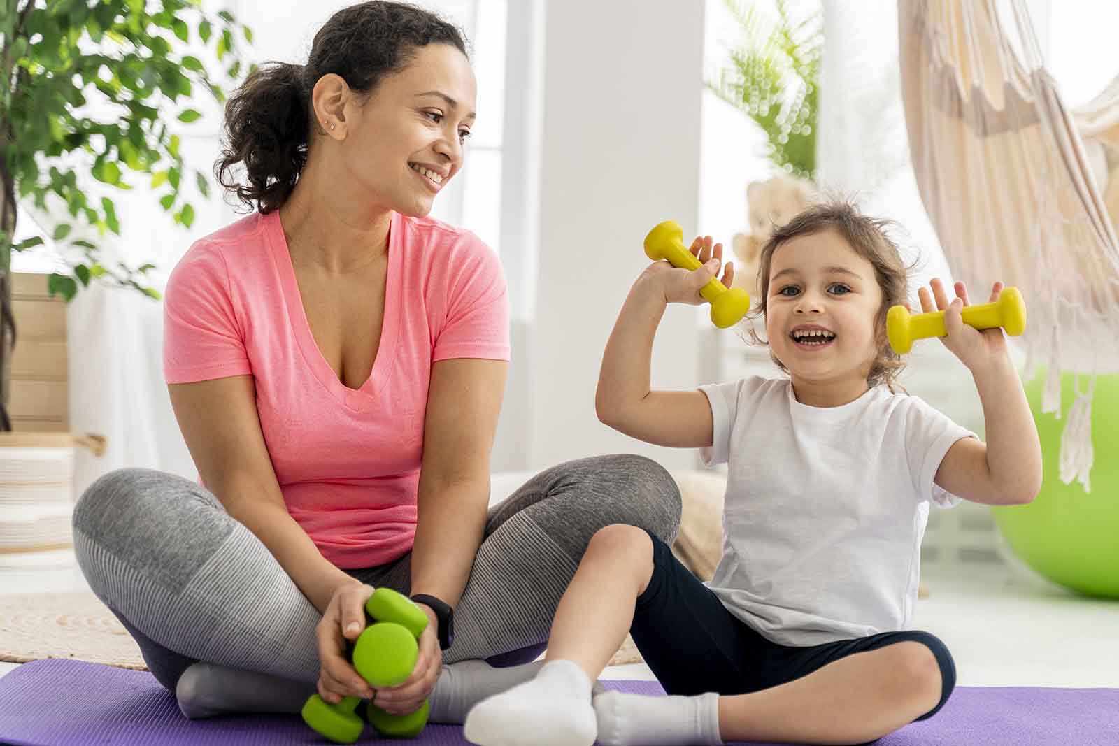 Imagem de uma menina e uma adulta sentadas em um tapete roxo no chão com roupas de praticar exercício, com a menina segurando pesos em suas mãos, com o fundo de sala de estar de dia