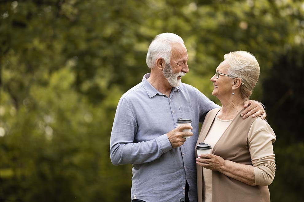 Imagem de um casal de idosos, juntos e se abraçando, cada um segurando um copo de café, com o fundo de árvores desfocadas e de dia. 