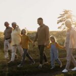 Imagem de uma família com avós, pais e crianças contentes andando juntas e de mãos dadas em um campo, durante o por do sol.