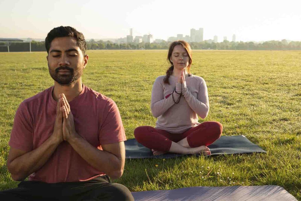 Imagem de uma dupla de modelos, vestindo roupas confortáveis, cada um sentados de pernas cruzadas sob um tapete, realizando uma meditação, em um gramado, durante o dia. 