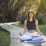 Imagem de uma modelo feminina, vestindo roupas de academia, sentada de pernas cruzadas em um tapete, realizando uma meditação, em um espaço com muitas árvores e grama ao redor e de dia.