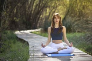 Imagem de uma modelo feminina, vestindo roupas de academia, sentada de pernas cruzadas em um tapete, realizando uma meditação, em um espaço com muitas árvores e grama ao redor e de dia.