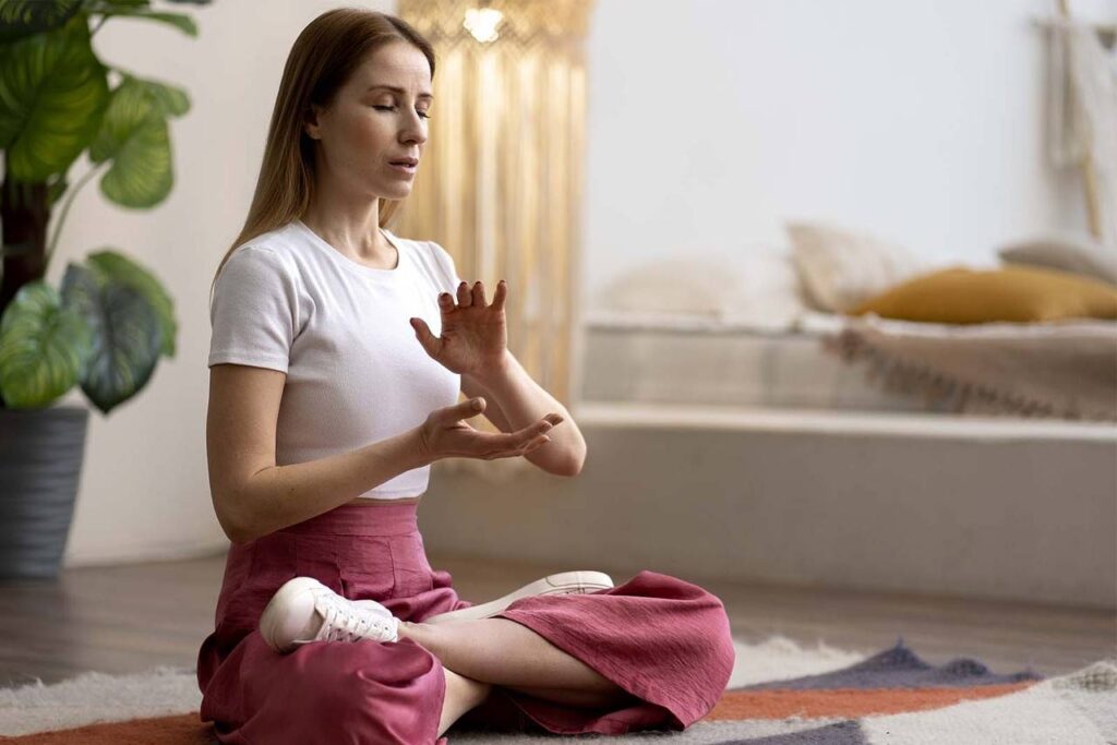 Imagem de uma modelo feminina, vestindo roupas confortáveis, sentada de pernas cruzadas sob um tapete, realizando uma meditação, no interior de uma sala, durante o dia. 