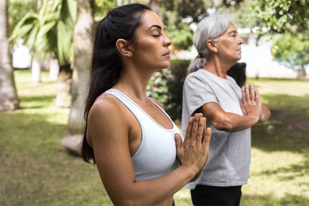 Imagem de uma dupla de modelos, uma com mais idade e a outra mais jovem, fazendo yoga ao ar livre de dia 